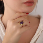 Close-up of a hand wearing a gold ring with a purple gemstone, against a neutral background.