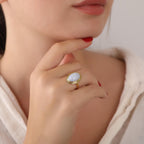Close-up of a hand wearing a gold ring with a blue lace agate gemstone, touching the chin.
