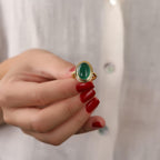 Hand wearing a gold ring with a green gemstone against a neutral background