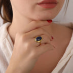 Close-up of a hand wearing a gold ring with a blue stone, touching the chin.
