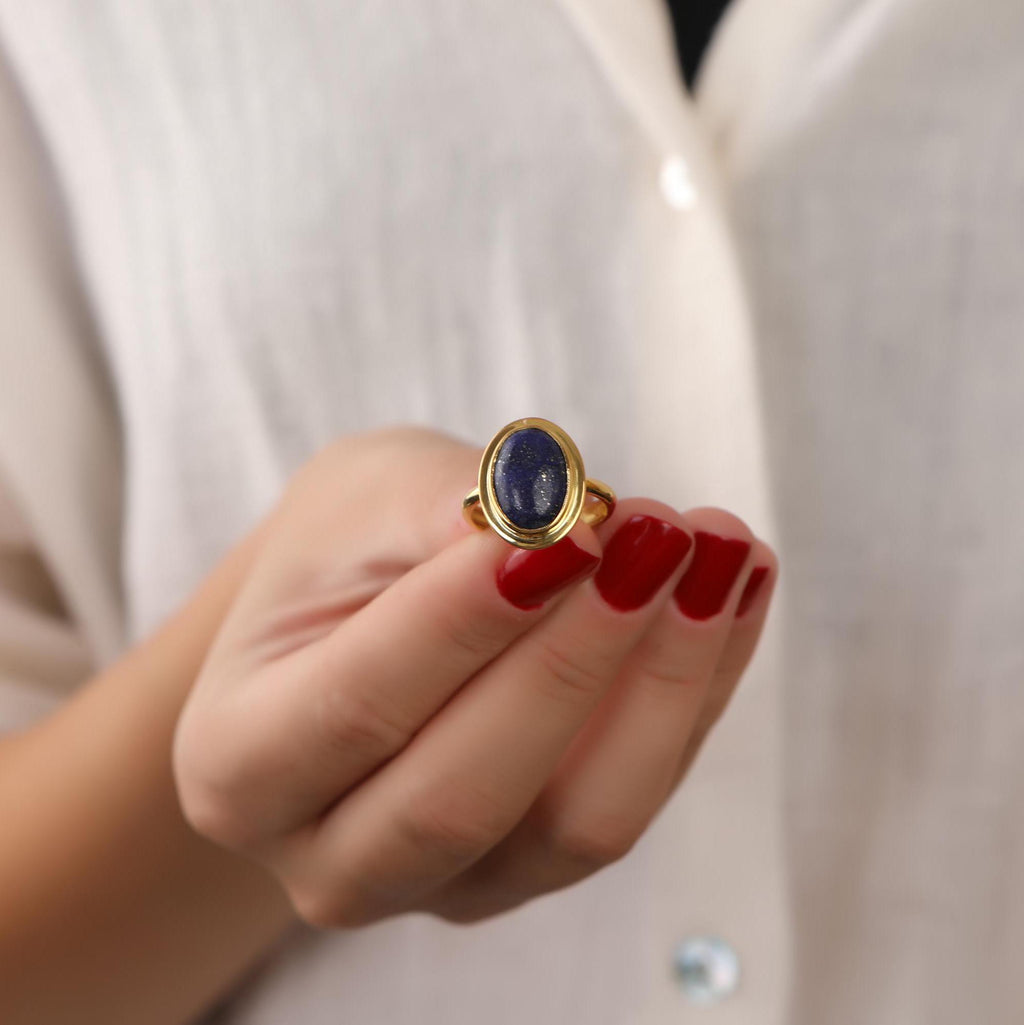 Hand wearing a gold ring with a blue stone against a neutral background