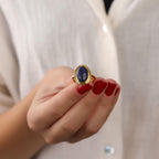 Hand wearing a gold ring with a blue stone against a neutral background