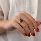 Hand wearing a gold ring with a blue stone against a neutral background