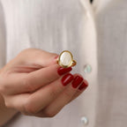 Hand wearing a gold ring with a white stone against a neutral background