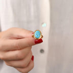 Gold ring with a turquoise stone held by a hand with red nail polish against a neutral background