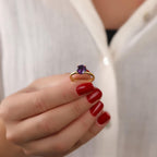 Gold ring with a purple gemstone on a hand with red nail polish against a white background