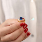 Gold ring with a blue gemstone held by a hand with red nail polish against a neutral background
