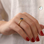 Hand wearing a gold ring with a blue gemstone against a neutral background