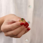 Gold ring with a green gemstone held by a hand with red nail polish against a blurred background
