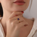 Close-up of a hand wearing a gold ring with a purple gemstone, touching the chin.