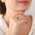 Close-up of a woman's hand wearing a gold ring with a green gemstone, touching her chin.