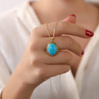 Hand holding a gold necklace with a turquoise pendant against a neutral background