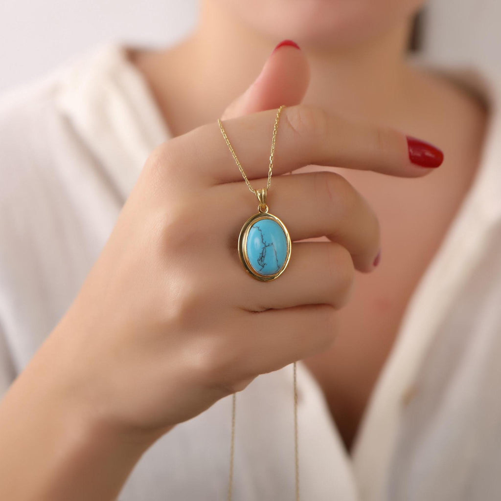 Hand holding a gold necklace with a turquoise pendant against a neutral background