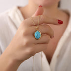 Hand holding a gold necklace with a turquoise pendant against a neutral background