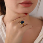 Close-up of a hand wearing a gold ring with a blue gemstone, touching the chin.
