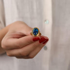 Hand wearing a gold ring with a blue gemstone against a light background