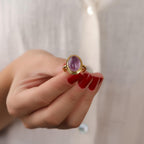 Hand wearing a gold ring with a purple gemstone against a neutral background