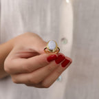 Hand wearing a gold ring with a large gemstone against a neutral background