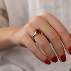 Hand wearing a gold ring with a red stone on a neutral background