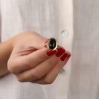 Hand wearing a gold ring with a black stone against a neutral background