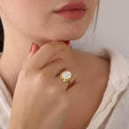 Close-up of a hand wearing a gold ring with a white stone, against a neutral background.