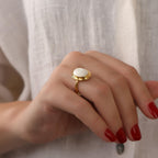 Hand wearing a gold ring with a white stone on a neutral background