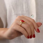 Hand wearing a gold ring with a red gemstone against a neutral background