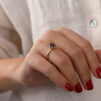 Hand wearing a gold ring with a purple gemstone against a neutral background