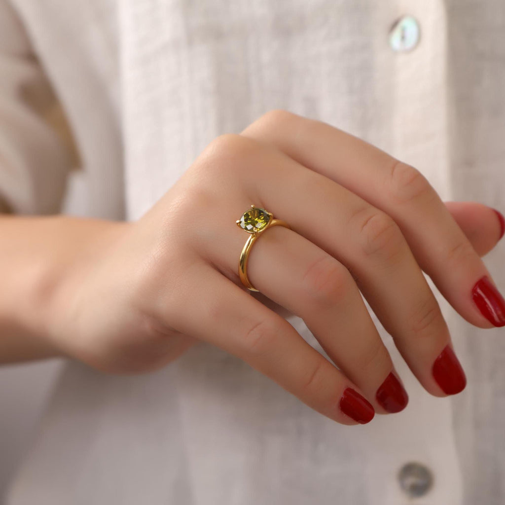 Hand wearing a gold ring with a green gemstone against a neutral background