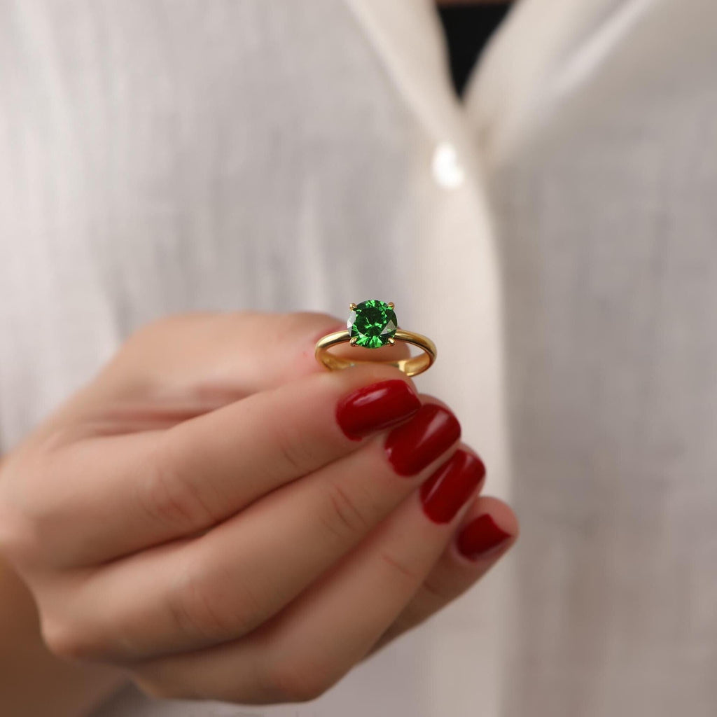 Gold ring with a green emerald gemstone held by a hand with red nail polish against a white background