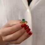 Gold ring with a green emerald gemstone held by a hand with red nail polish against a white background