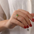 Hand wearing a gold ring with a green gemstone against a neutral background