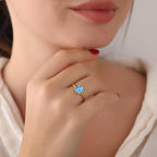 Close-up of a woman's hand wearing a gold ring with a blue gemstone, blurred background.