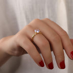 Hand wearing a gold ring with a purple gemstone on a neutral background