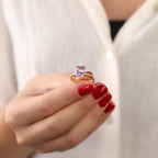 Hand with red nail polish holding a gold ring with a purple gemstone against a white background