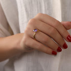 Hand wearing a gold ring with a purple gemstone against a neutral background