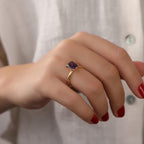 Hand wearing a gold solitaire ring with a purple gemstone against a neutral background