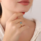 Close-up of a hand wearing a gold ring with a blue gemstone, touching the chin.