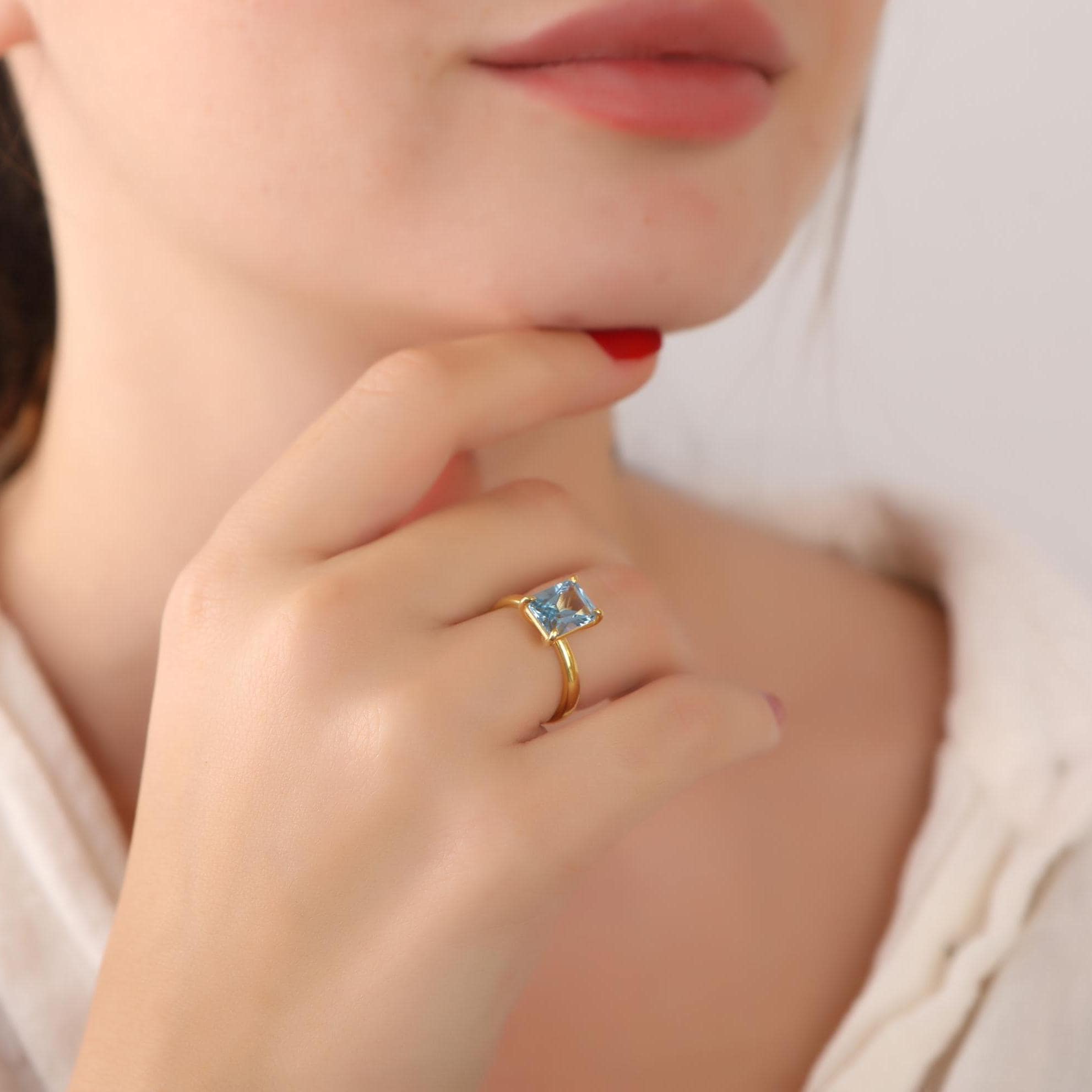 Close-up of a hand wearing a gold ring with a blue gemstone, touching the chin.