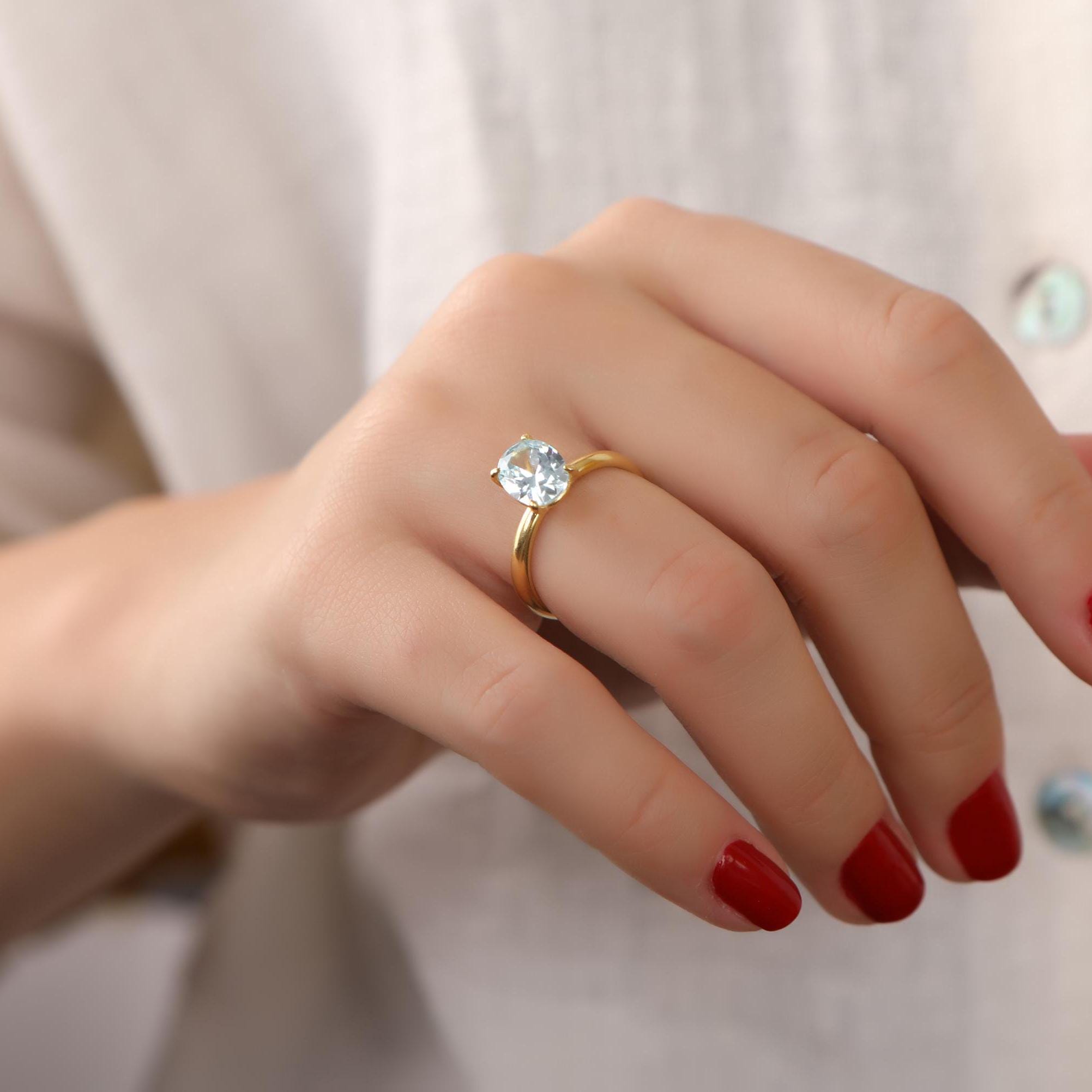 Hand wearing a gold ring with a clear gemstone on a neutral background