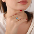 Close-up of a hand wearing a gold ring with a blue gemstone, touching the chin.