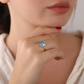 Close-up of a hand wearing a gold ring with a rectangular blue gemstone, held near the face.