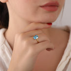 Close-up of a hand wearing a gold ring with a rectangular blue gemstone, held near the face.