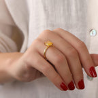 Hand wearing a gold ring with a yellow stone against a neutral background