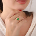 Close-up of a hand wearing a gold ring with a green gemstone, against a blurred background.