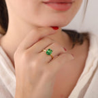 Close-up of a hand wearing a gold ring with a green gemstone, against a blurred background.