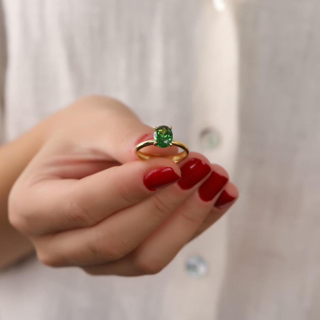 Hand wearing a gold ring with a green gemstone against a neutral background