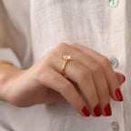 Hand wearing a gold solitaire ring with red nail polish on a neutral background
