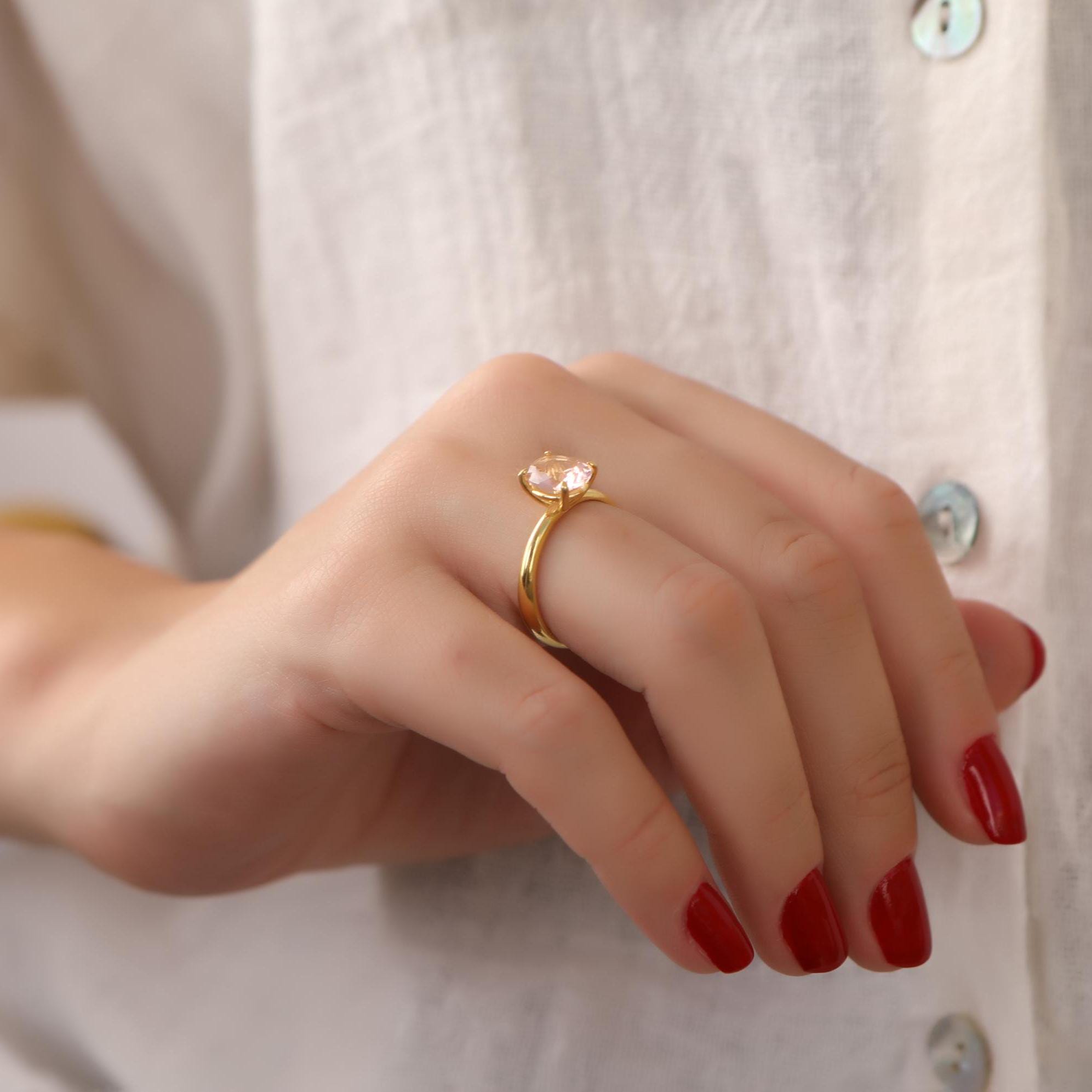 Hand wearing a gold solitaire ring with red nail polish on a neutral background