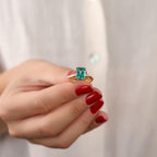Hand with red nail polish holding a ring with a green gemstone against a plain background